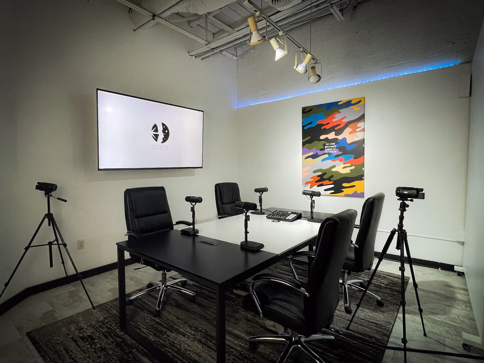CREATRVERSE podcast and media studio with black conference table, black executive chairs, multiple cameras on tripods, wall-mounted screen showing Creatr logo, colorful camo-print motivational poster, track lighting, and blue LED accent strip along ceiling