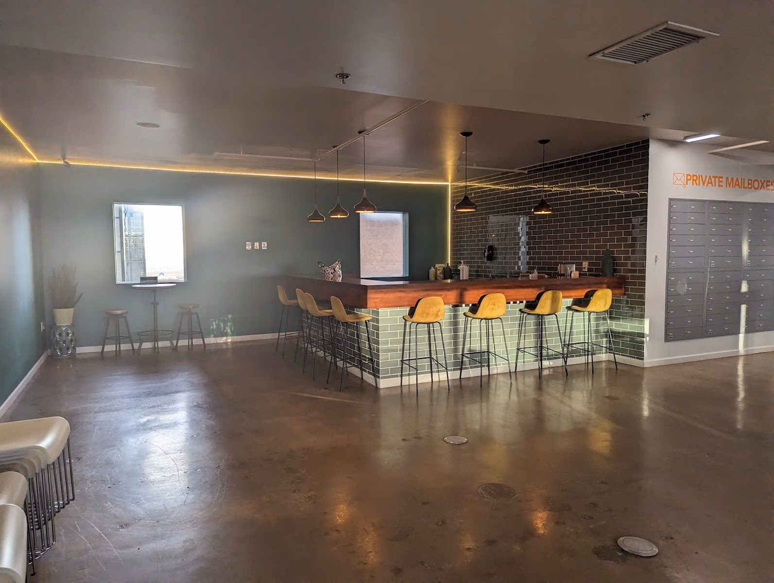 Connected Coworking moody bar-style common area with sage green walls, dark brick accent wall, walnut-topped L-shaped bar counter with green subway tile base, mustard yellow Eames-style bar stools, black pendant lights, private mailbox wall with orange lettering, and polished concrete floors