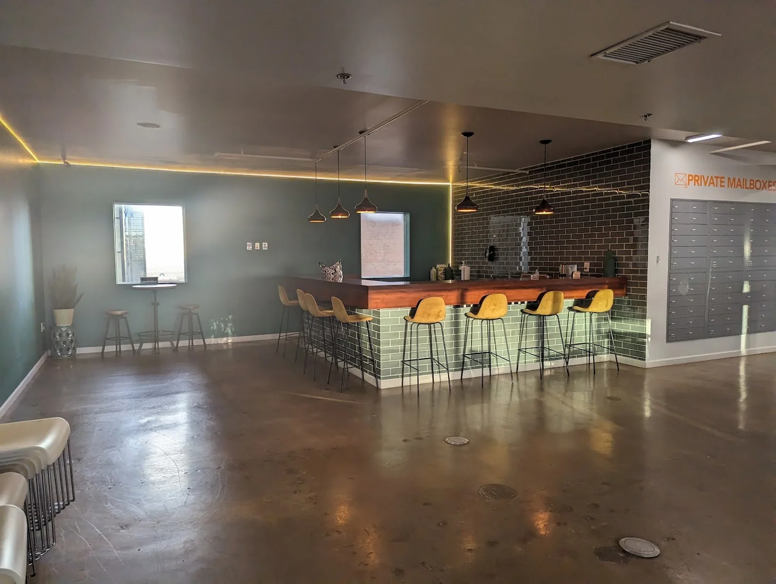 Connected Coworking moody bar-style common area with sage green walls, dark brick accent wall, walnut-topped L-shaped bar counter with green subway tile base, mustard yellow Eames-style bar stools, black pendant lights, private mailbox wall with orange lettering, and polished concrete floors