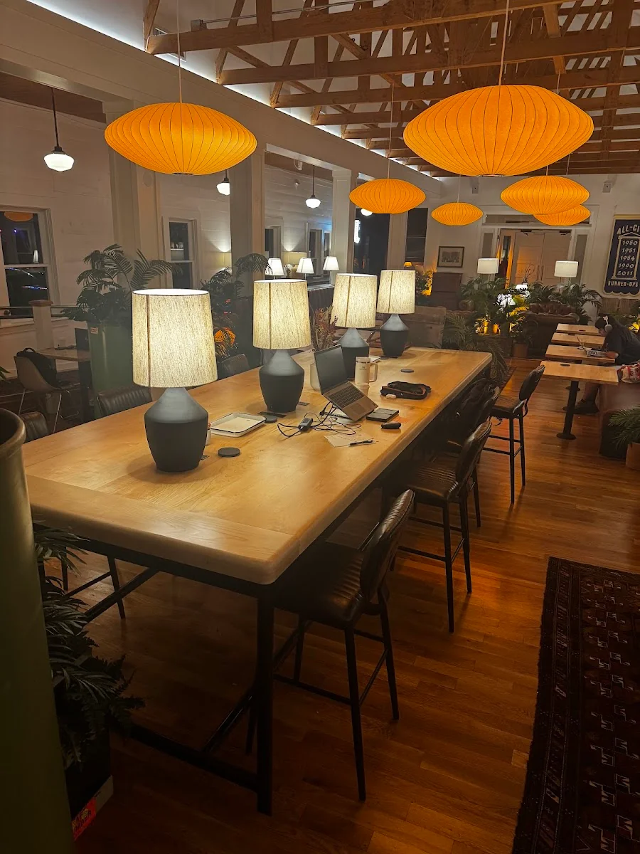 Switchyards Dallas warm evening-lit coworking space in converted house with exposed timber rafters, large orange George Nelson-style bubble pendant lamps, long oak communal table with ceramic table lamps, dark leather chairs, hardwood floors, and lush tropical plants