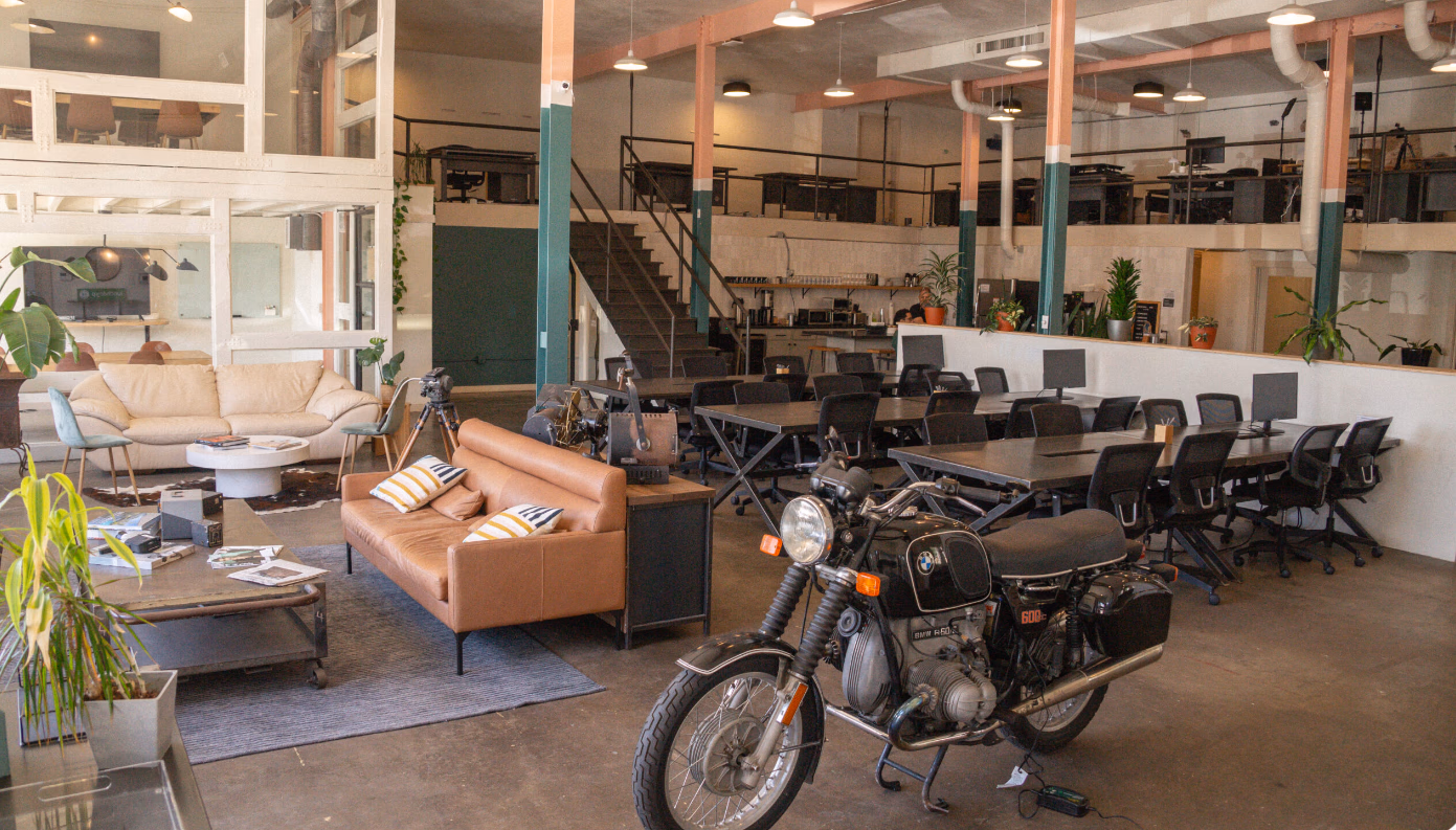 E-Creative Space eclectic double-height warehouse with vintage BMW motorcycle as decor centerpiece, tan leather and cream sofas on cowhide rug, teal-and-salmon painted steel columns, mezzanine level with desk stations, metal staircase, coffee bar, and potted plants throughout