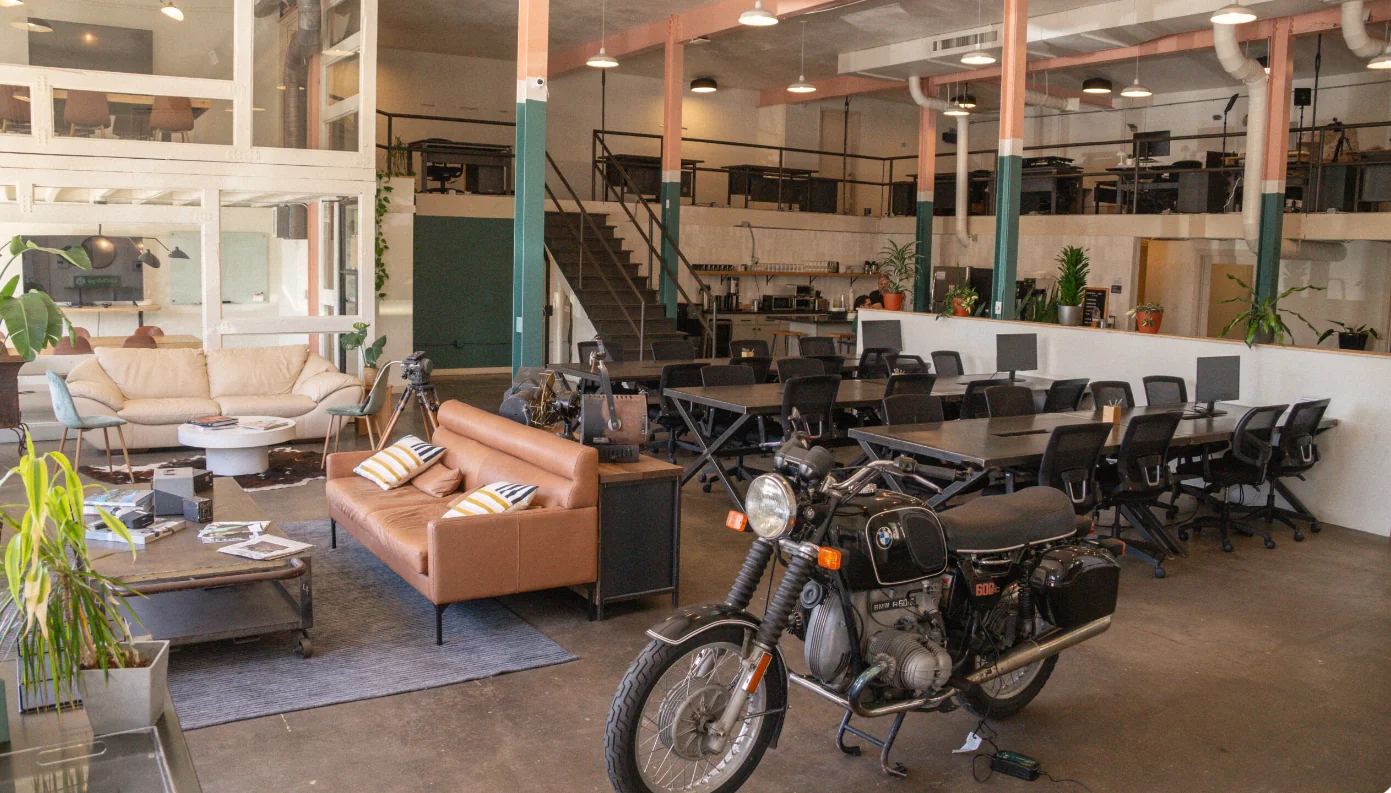 E-Creative Space eclectic double-height warehouse with vintage BMW motorcycle as decor centerpiece, tan leather and cream sofas on cowhide rug, teal-and-salmon painted steel columns, mezzanine level with desk stations, metal staircase, coffee bar, and potted plants throughout