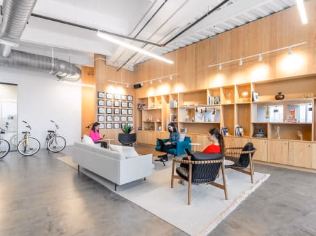 Spaces Deep Ellum modern lounge with warm wood built-in shelving unit displaying books and decor, white sofa, teal accent chair, woven black lounge chair, three women chatting, bicycles parked by wall, exposed ductwork, polished concrete floor, and track lighting