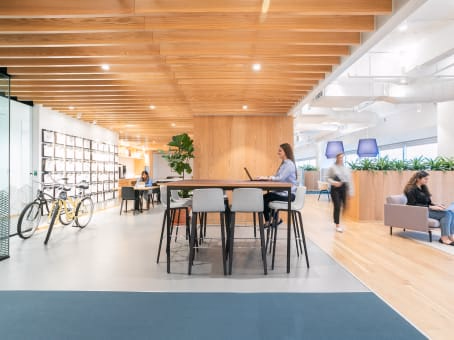 Spaces The Colonnade bright open workspace with warm wood slat ceiling, high communal table with black chairs, woman standing with laptop, bicycles parked near display shelving wall, fiddle-leaf fig plant, light oak floors, planter dividers, and members working on grey sofas