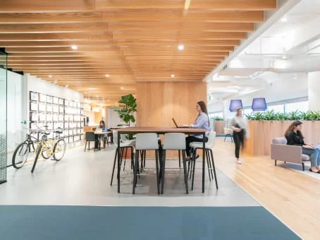 Spaces The Colonnade bright open workspace with warm wood slat ceiling, high communal table with black chairs, woman standing with laptop, bicycles parked near display shelving wall, fiddle-leaf fig plant, light oak floors, planter dividers, and members working on grey sofas