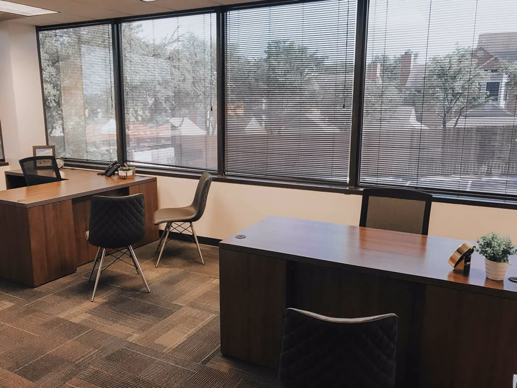 ExecutiveWorkspace Hillcrest Green private office with two walnut L-shaped desks, quilted grey and brown accent chairs, large windows with mini-blinds showing tree-lined views, patterned carpet tiles, desk phone, and small plant