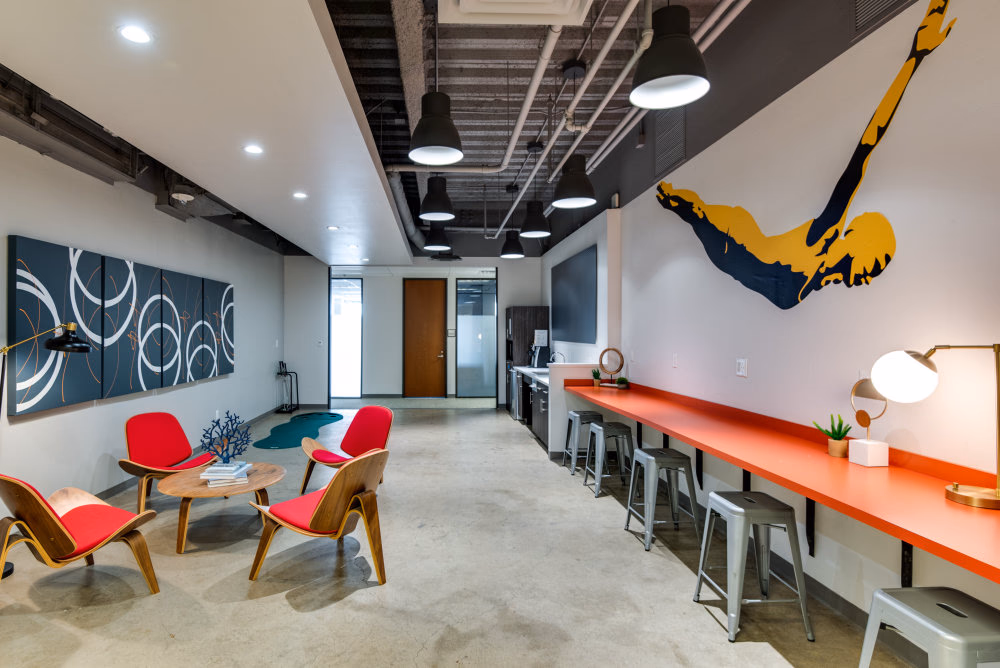 Workstyle Spaces Pacific Ave industrial lounge with red mid-century shell chairs around a small wood table, navy abstract swirl art panels on grey wall, long orange counter bar with metal Tolix stools, bold yellow-and-navy diver wall mural, exposed ceiling with black pendant lights, and polished concrete floors