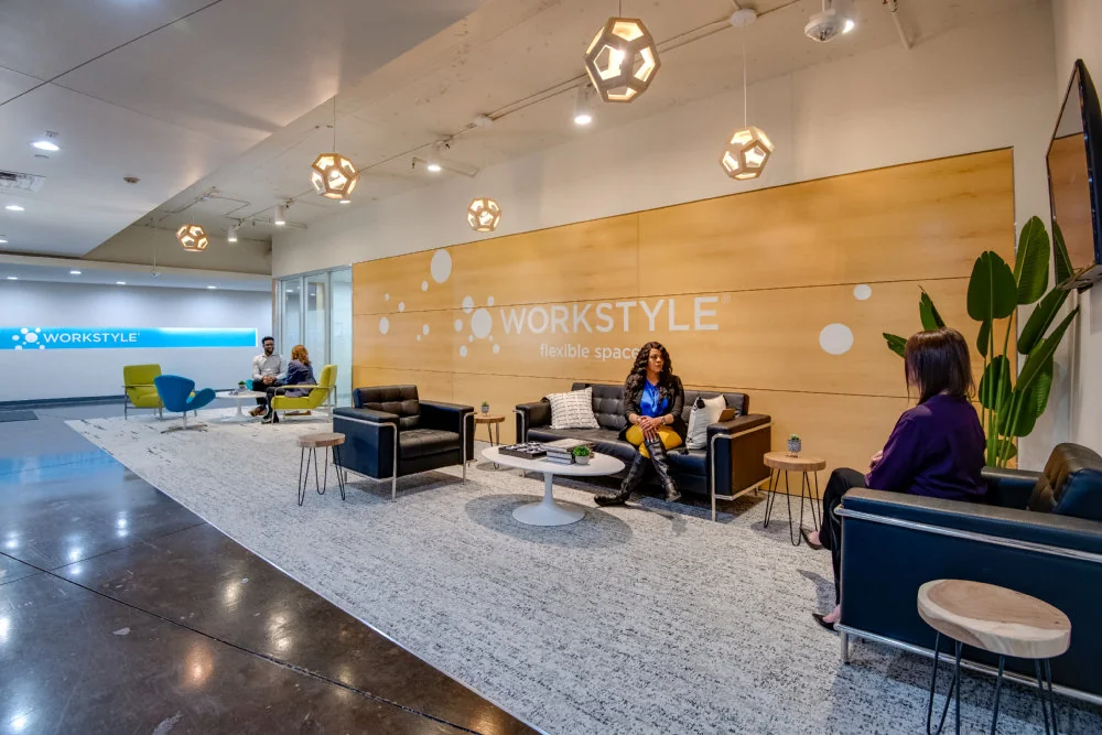 Workstyle Spaces North Central Expressway spacious lobby with large wood panel feature wall displaying Workstyle flexible space branding, dark leather sofas and club chairs, white tulip coffee table, geometric gold pendant lights, polished concrete floors, green accent chairs in background, and bird-of-paradise plant