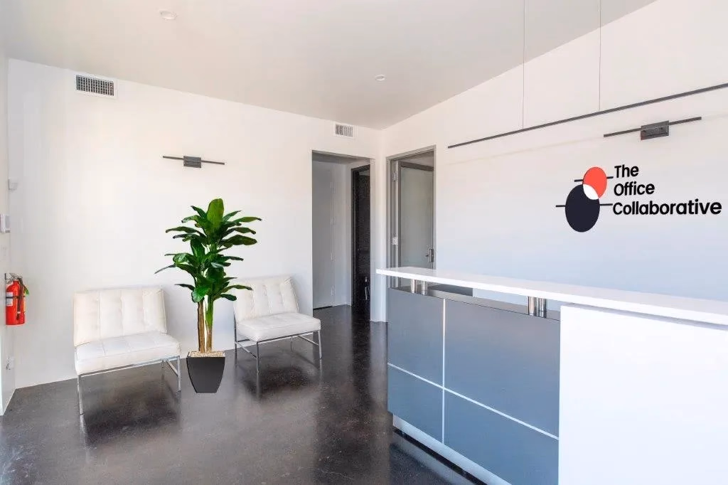 The Office Collaborative minimalist white reception area with red-and-black logo on wall, white Barcelona-style lounge chairs, tropical green plant, sleek grey reception desk with white countertop, dark polished concrete floors, and clean modern picture lights