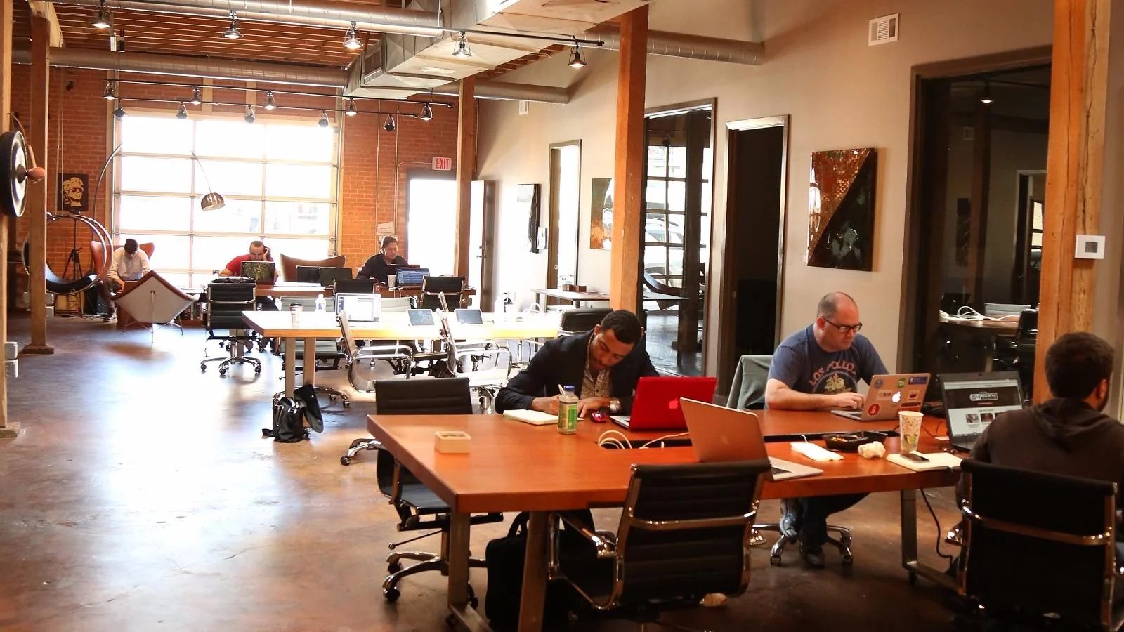GeniusDen raw industrial warehouse workspace with exposed brick walls, timber columns, roll-up garage door flooding natural light, polished concrete floors, large wooden communal tables where members work on laptops, track lighting, and glass-walled private offices
