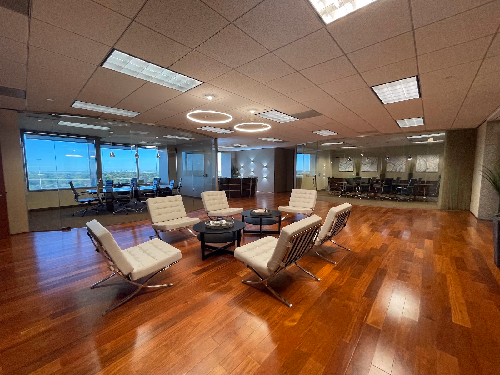 Lucid Private Offices Galleria executive lobby with rich cherry hardwood floors, white Barcelona chairs arranged around a black coffee table, glass-walled conference rooms on both sides, ring pendant lights, and floor-to-ceiling windows with panoramic views