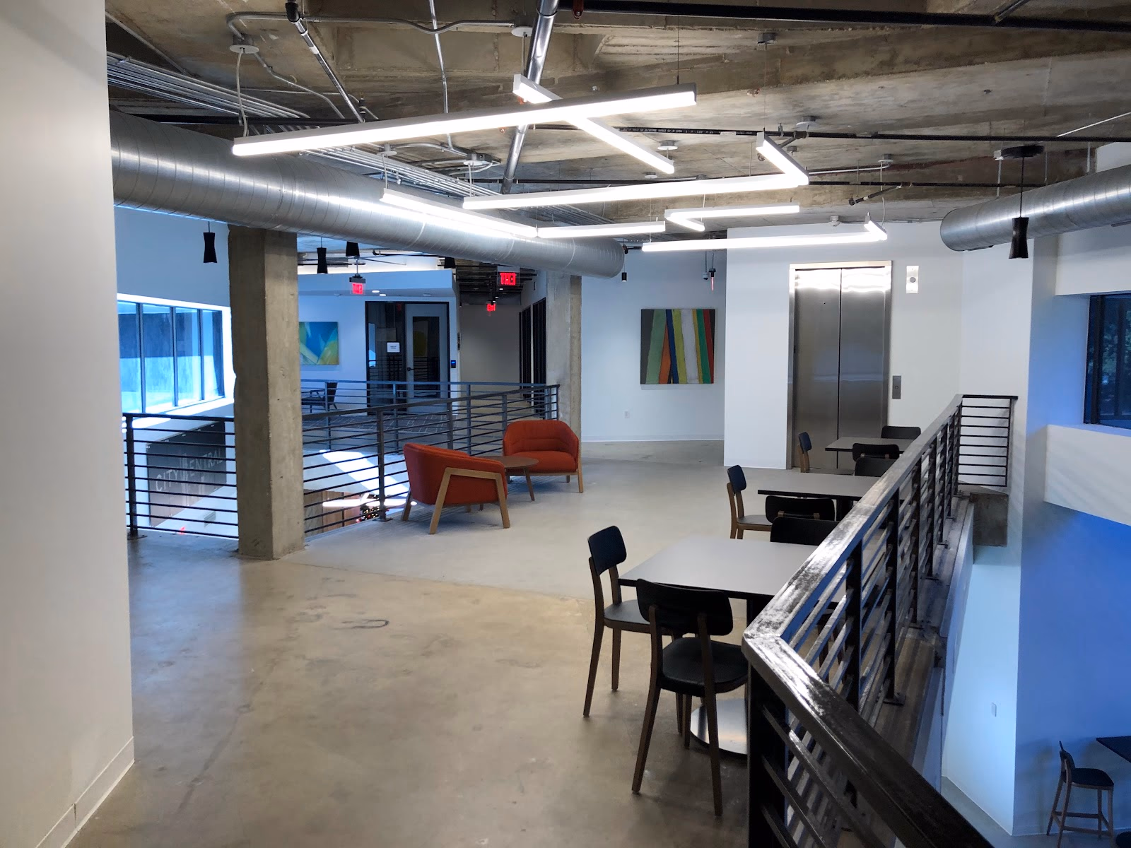 CityCentral North Dallas industrial-modern mezzanine level with polished concrete floors, exposed concrete columns and ceiling, geometric LED light fixtures, metal railings, scattered black chairs and tables, a red accent armchair, and colorful striped art by the elevator