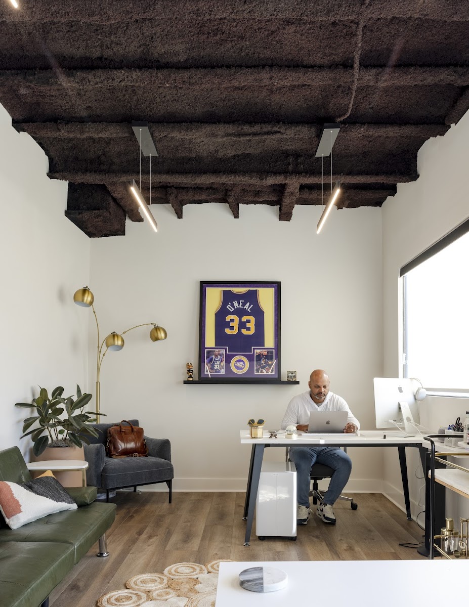 The Rigs Workspace private office with framed Shaq O'Neal jersey, exposed dark ceiling beams, brass floor lamp, and member at dual-monitor desk