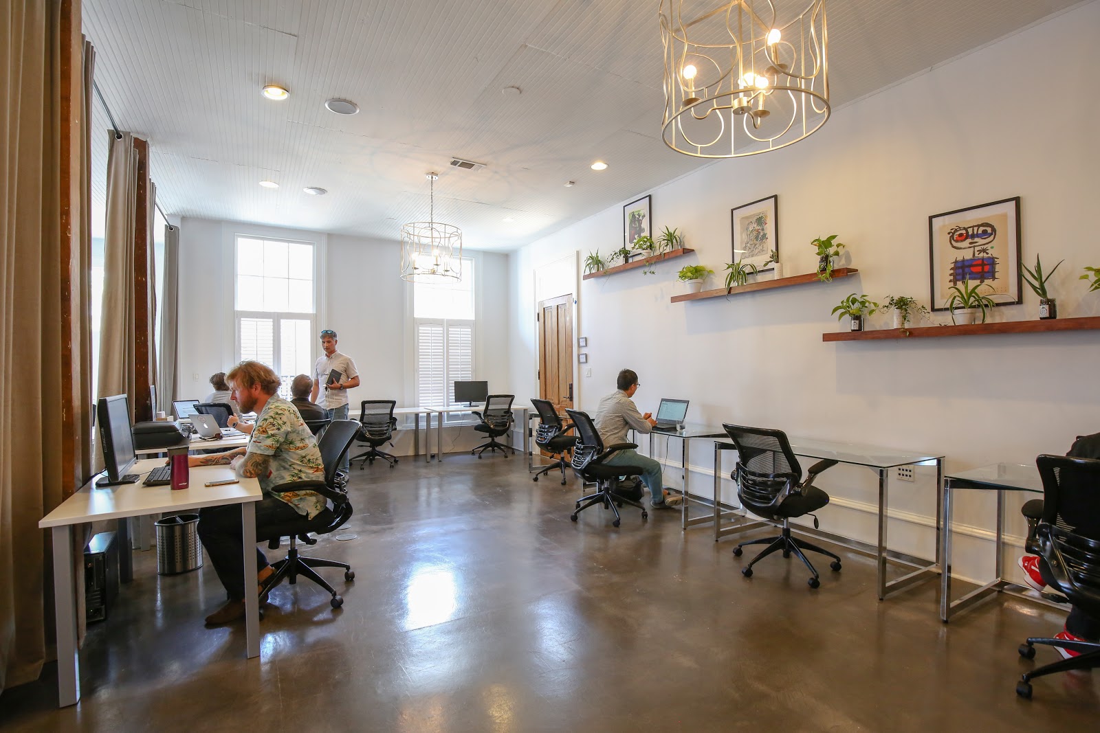 UrbanHUB LGD bright coworking room with white beadboard ceiling, ornate gold chandelier, polished concrete floors, and plant shelves with NOLA art