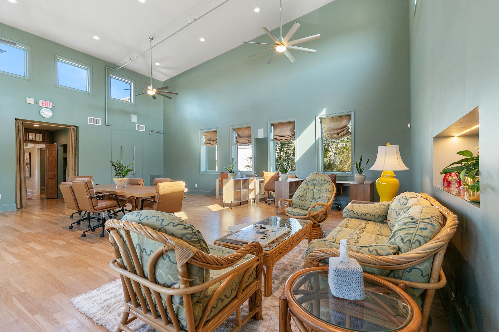 The CoWorking Studio of Jena NOLA vaulted sage-green room with rattan furniture, wave-patterned cushions, ceiling fans, and yellow ceramic lamp