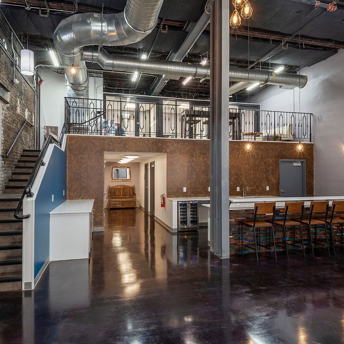 WB Collective industrial two-story space with polished dark concrete floors, wrought-iron mezzanine railing, Edison bulb pendants, and reclaimed-wood bar