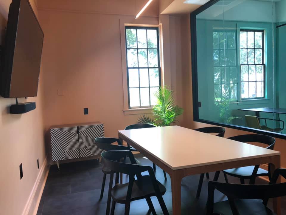 BayouWorks meeting room with salmon walls, white conference table, black chairs, geometric credenza, wall-mounted TV, and glass partition