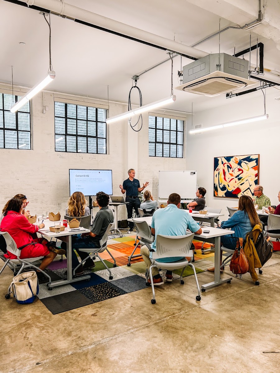 Scale New Orleans warehouse meeting space with white brick walls, colorful patchwork rug, presenter giving Cohort 6 HQ talk, and abstract art