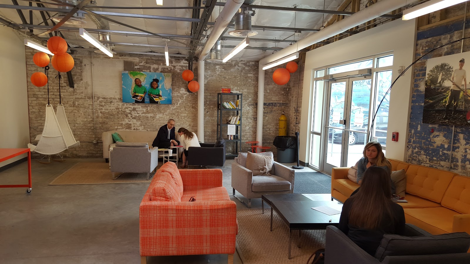 Propeller incubator lounge with exposed brick, orange plaid sofa, gray armchairs, orange paper lanterns, hammock chair, and colorful art