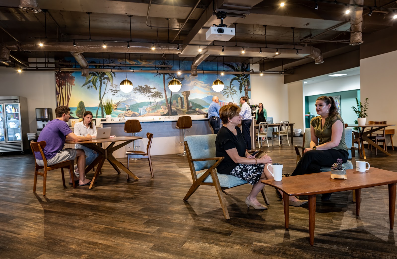 Connect Hub Coworking with tropical mural backdrop, wood floors, mid-century blue chairs, brass pendant lights, and members chatting