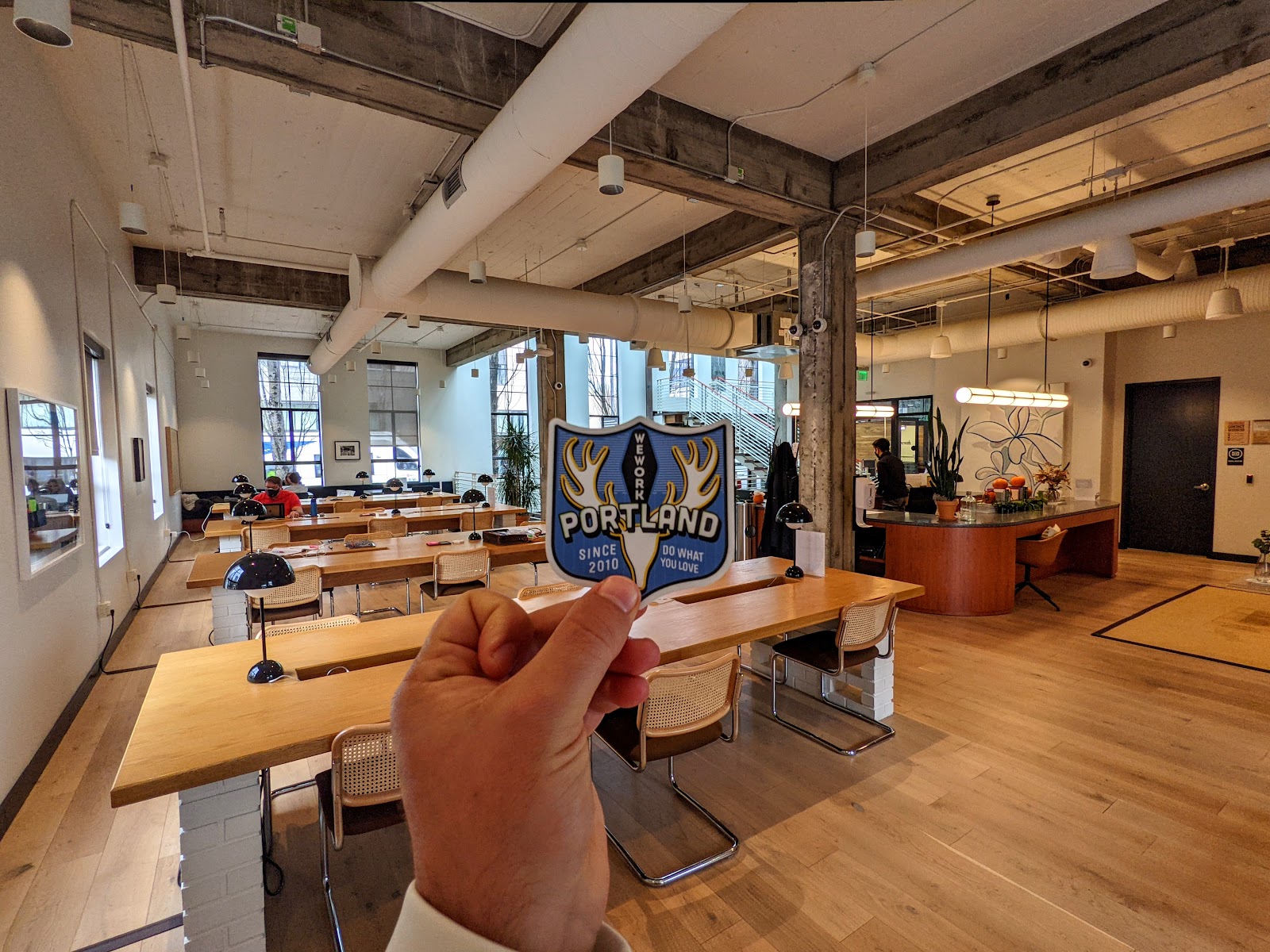 WeWork Portland open workspace with birch communal tables, cane-back Cesca chairs, concrete beams, hardwood floors, and hand holding a WeWork Portland sticker