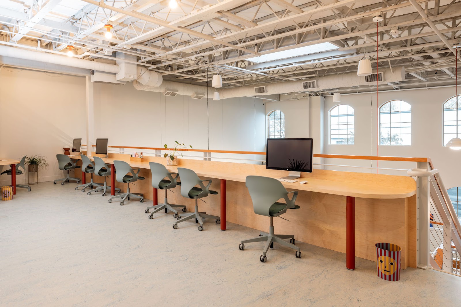 It's Just a Feeling bright white studio with exposed steel trusses, arched windows, long curved birch plywood communal desk with red legs, and sage-green task chairs