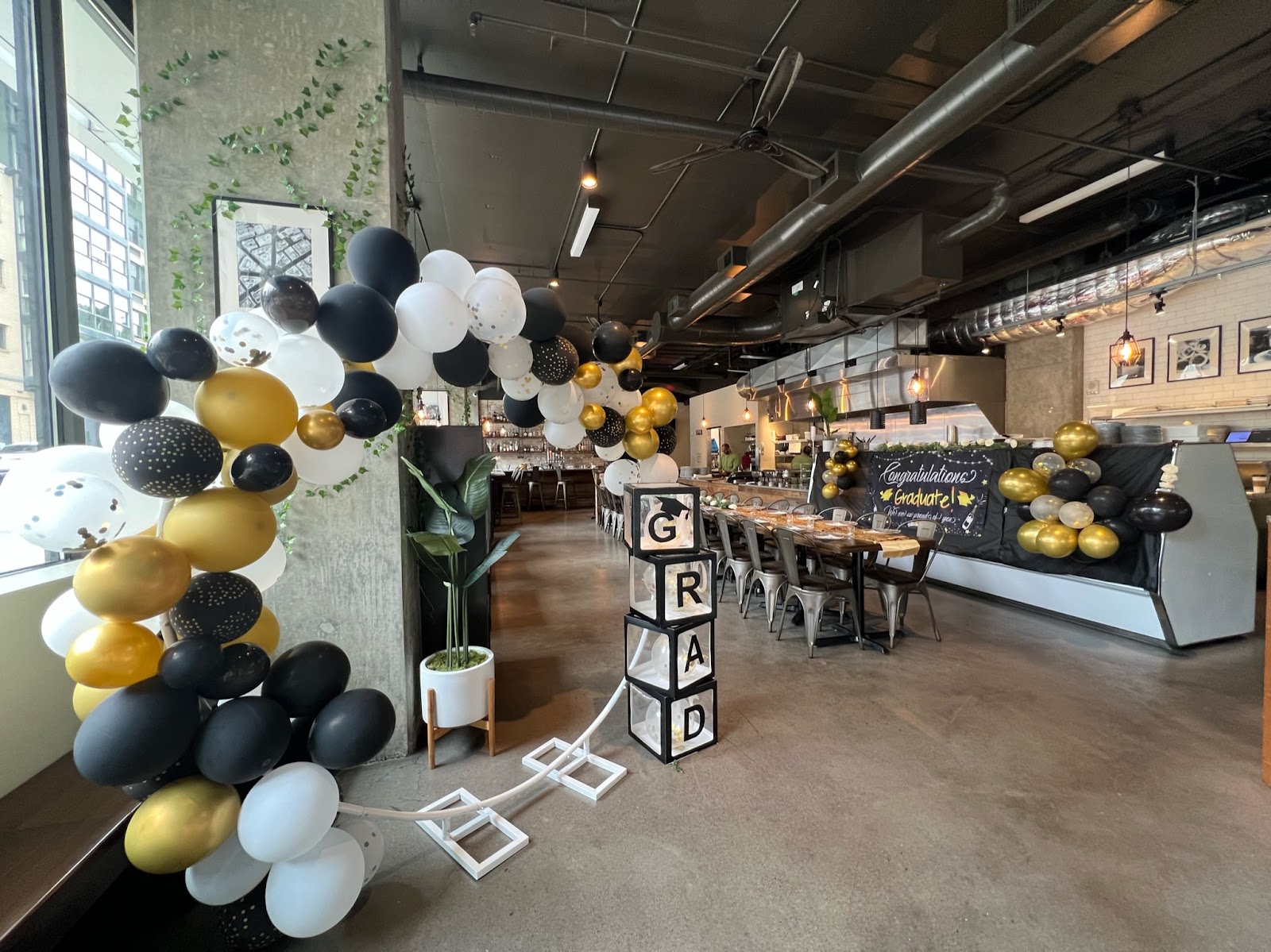 The Penthouse by KaramPDX event space with black-gold-white balloon arch, GRAD letter blocks, long communal wood table, metal chairs, exposed concrete columns