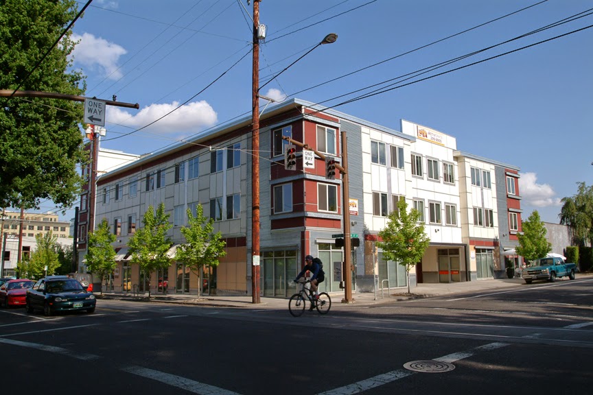ActivSpace on Lovejoy modern three-story mixed-use building with gray and burgundy metal cladding, street-level storefronts, cyclist passing, and green trees