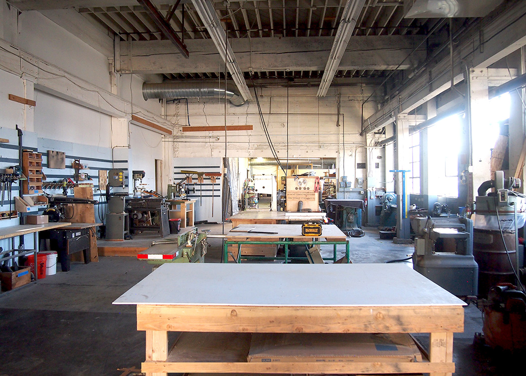 Past Lives Makerspace industrial woodworking shop with large work tables, band saws, drill presses, power tools, high ceilings with exposed beams, and natural light