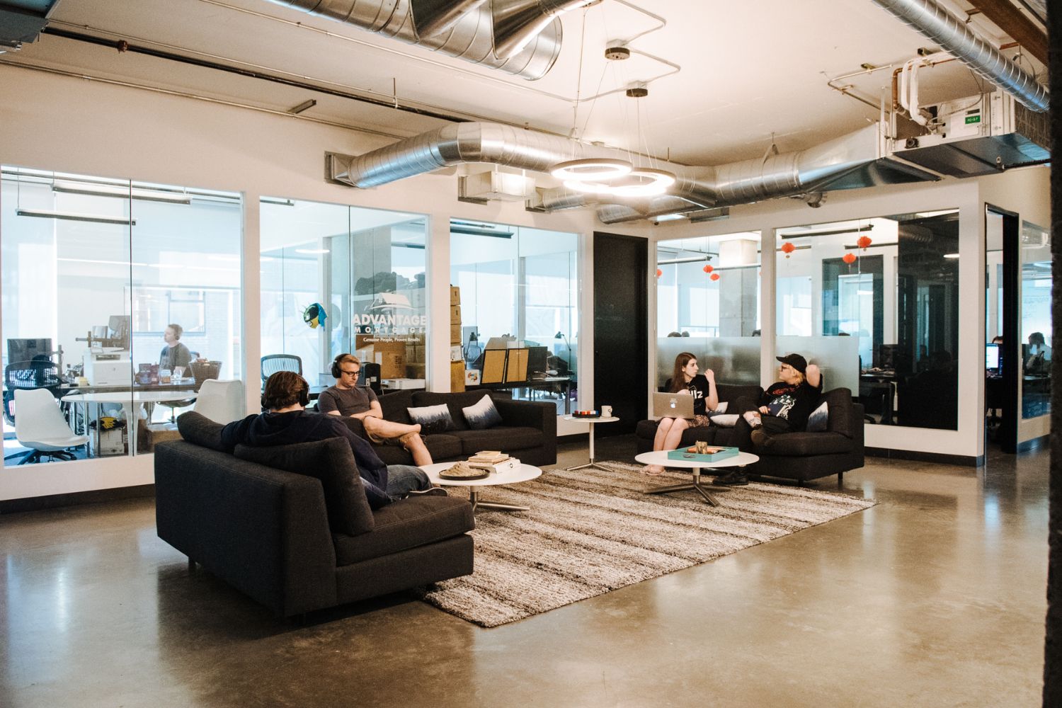CENTRL Office Eastside open lounge with dark gray sectional sofas, striped area rug, white coffee tables, exposed HVAC ducts, and glass-walled private offices