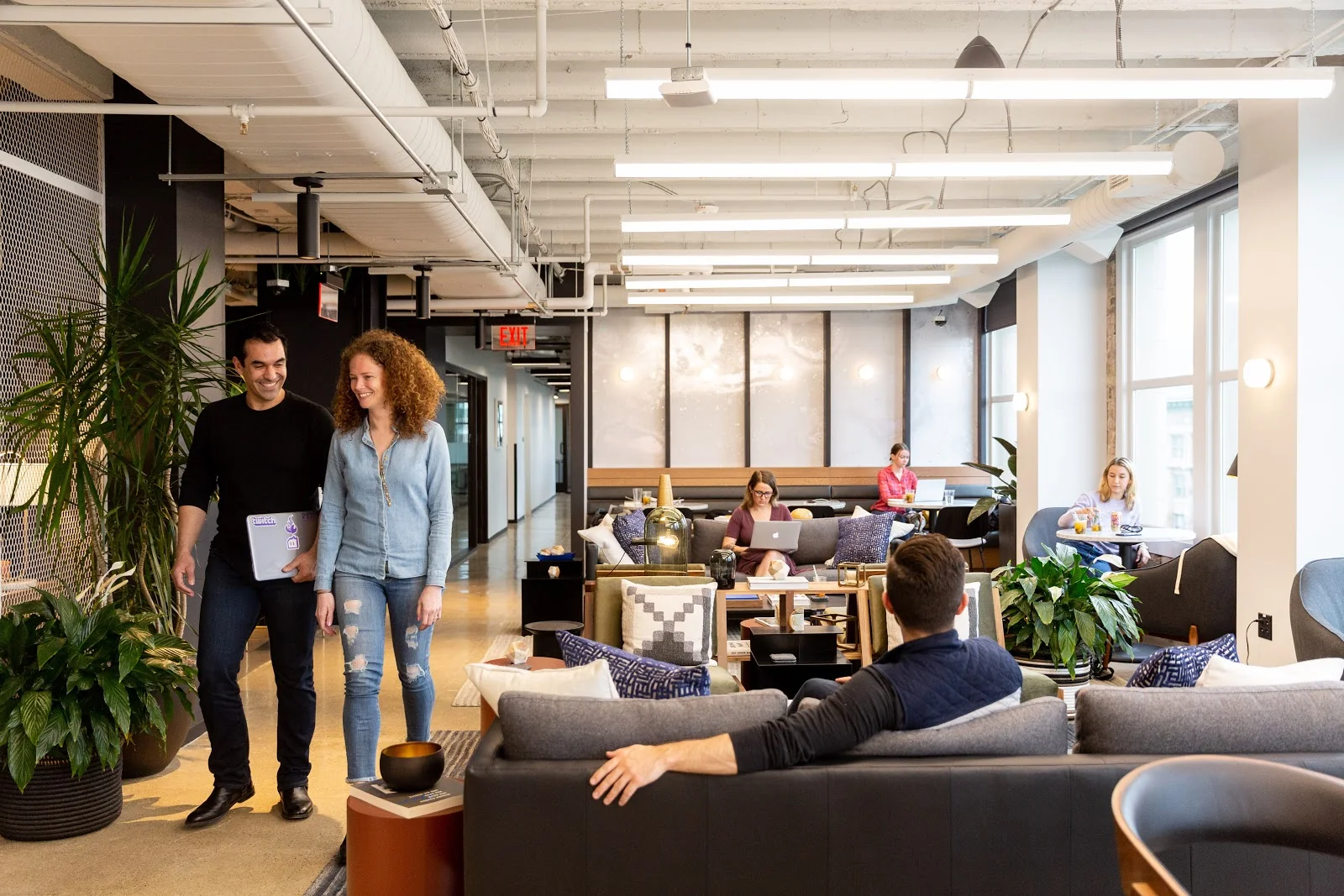 Industrious Portland polished lounge with gray sofas, patterned throw pillows, potted plants, members chatting and working on laptops by large windows