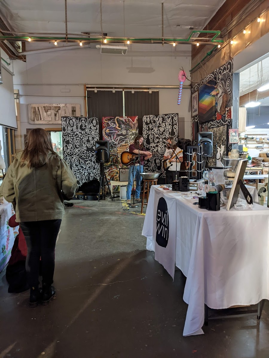 ADX Art Design Xchange warehouse art market with live musicians performing in front of psychedelic murals, vendor tables, string lights, and METAL sign