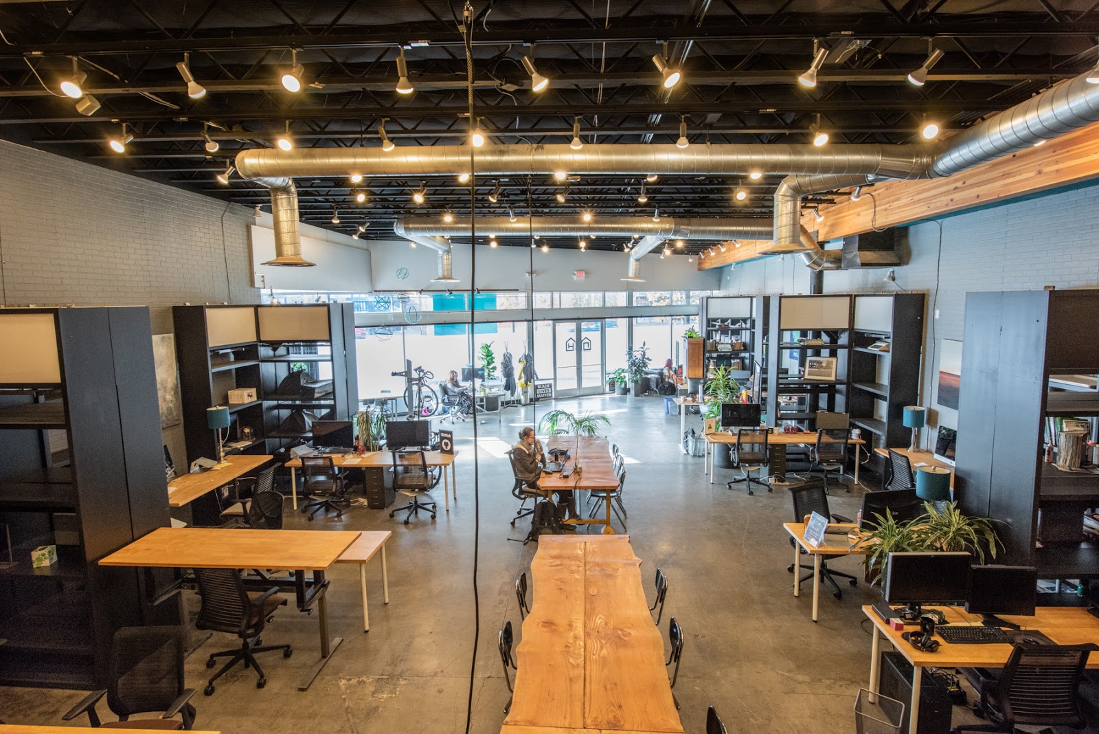 openHAUS coworking warehouse floor with black ceiling, track lighting, live-edge wood communal table, individual desks with privacy partitions, plants, and polished concrete