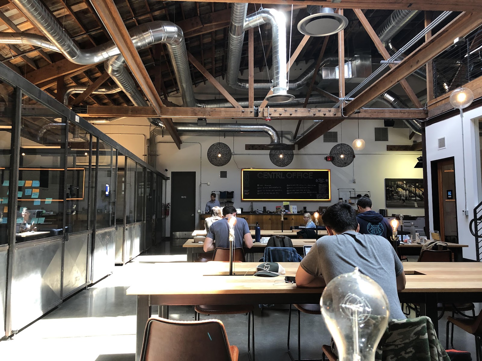 CENTRL Office Pearl industrial interior with exposed wood-beam ceiling, HVAC ducts, CENTRL OFFICE chalkboard menu, butcher-block communal tables, and members working