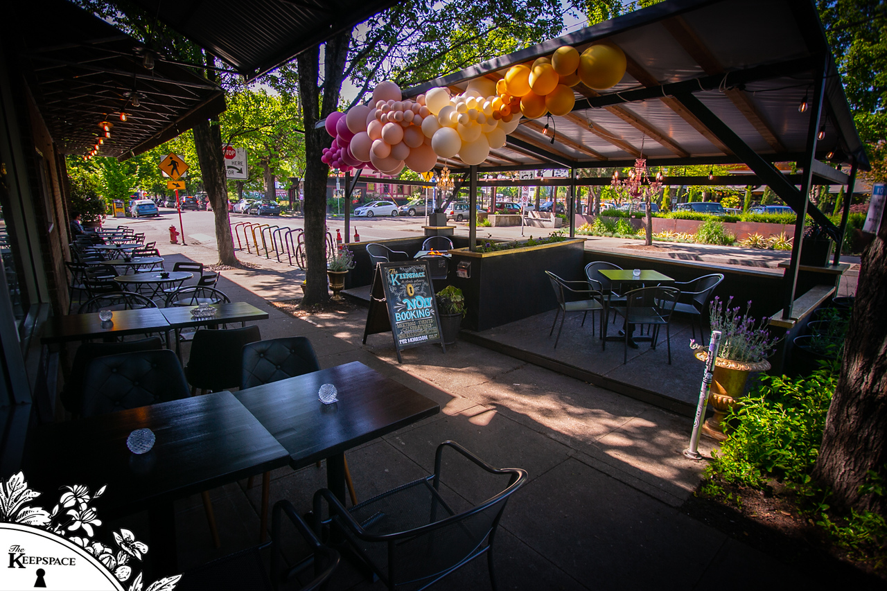 The Keepspace covered outdoor patio with black metal tables, colorful pink-gold-cream balloon garland, chalkboard A-frame sign, and tree-lined SE Portland street