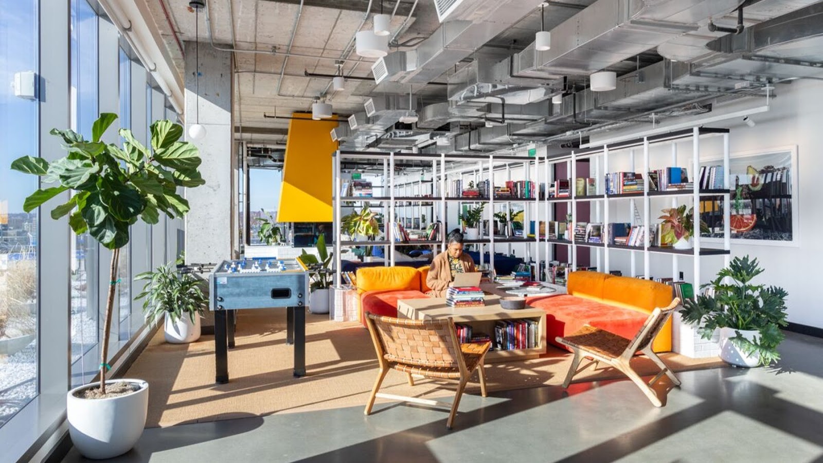 WeWork vibrant lounge with orange and yellow curved sofa, foosball table, bookshelves, large plants, and natural light