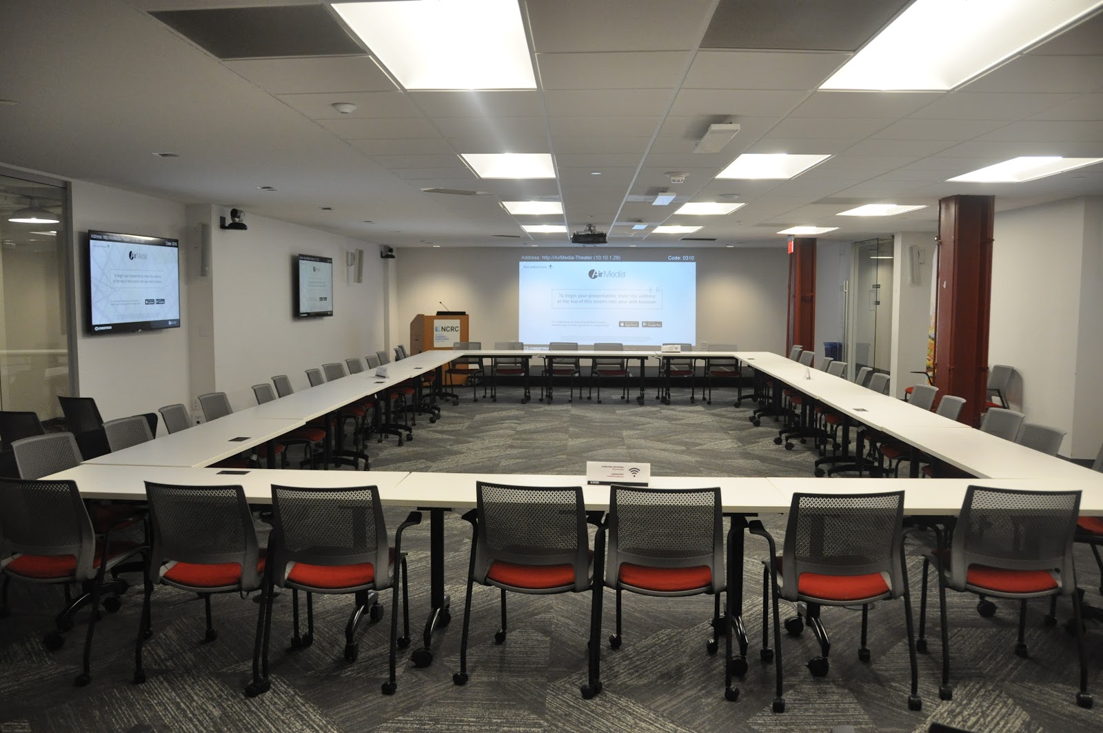 Just Economy Club large U-shaped conference setup with white tables, red-cushioned mesh chairs, projector screen, and dual TV monitors